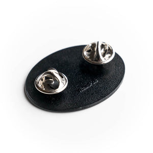 Black round badge with two silver brooches on a white background